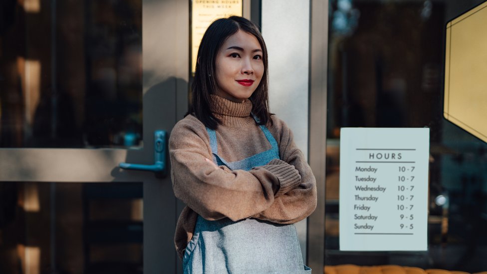 Mulher sorrindo de forma contida em frente a estabelecimento, aparentemente um café