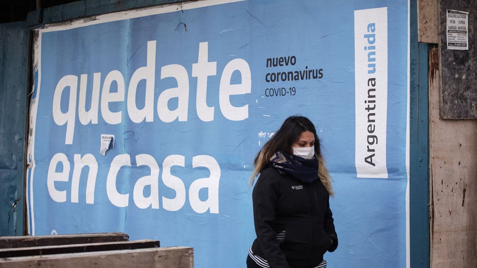 Mulher com máscara passa em frente a cartaz que diz "fique em casa" em espanhol