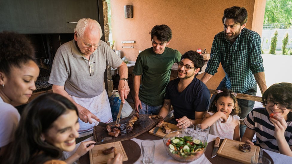 Adultos, adolescentes e crianças se reúnem em volta de uma mesa, onde um homem idoso corta uma carne de churrasco