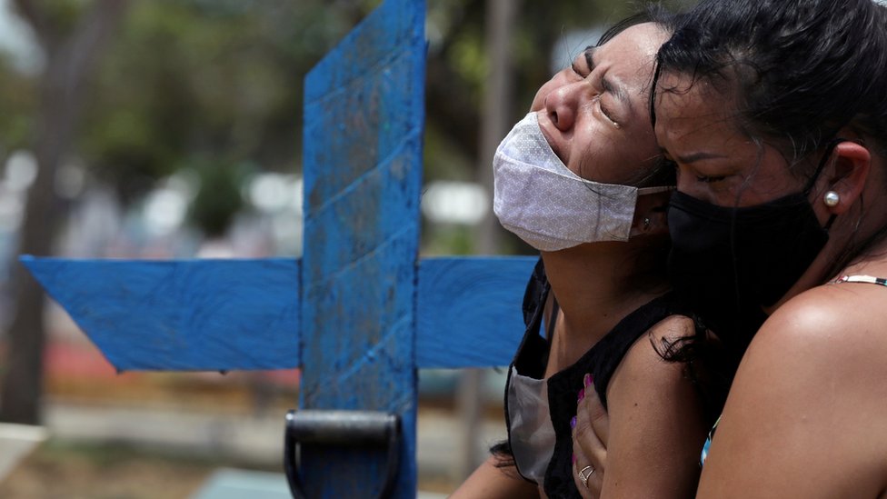 Kelvia Andrea Goncalves, 16, uma jovem de cabelos lisos e pretos, de máscara, chora de olhos fechados e com expressão de grande dor, e é abraçada pela tia, uma mulher de meia idade e cabelos pretos, em frente a uma cruz de madeira azul que marca o túmulo da mãe, no cemitério Parque Taruma, em Manaus