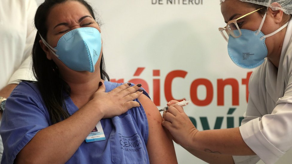 Mulher de cabelo preto comprido, pela parda e uniforme de profissional de saúde recebe vacina no Rio de Janeiro