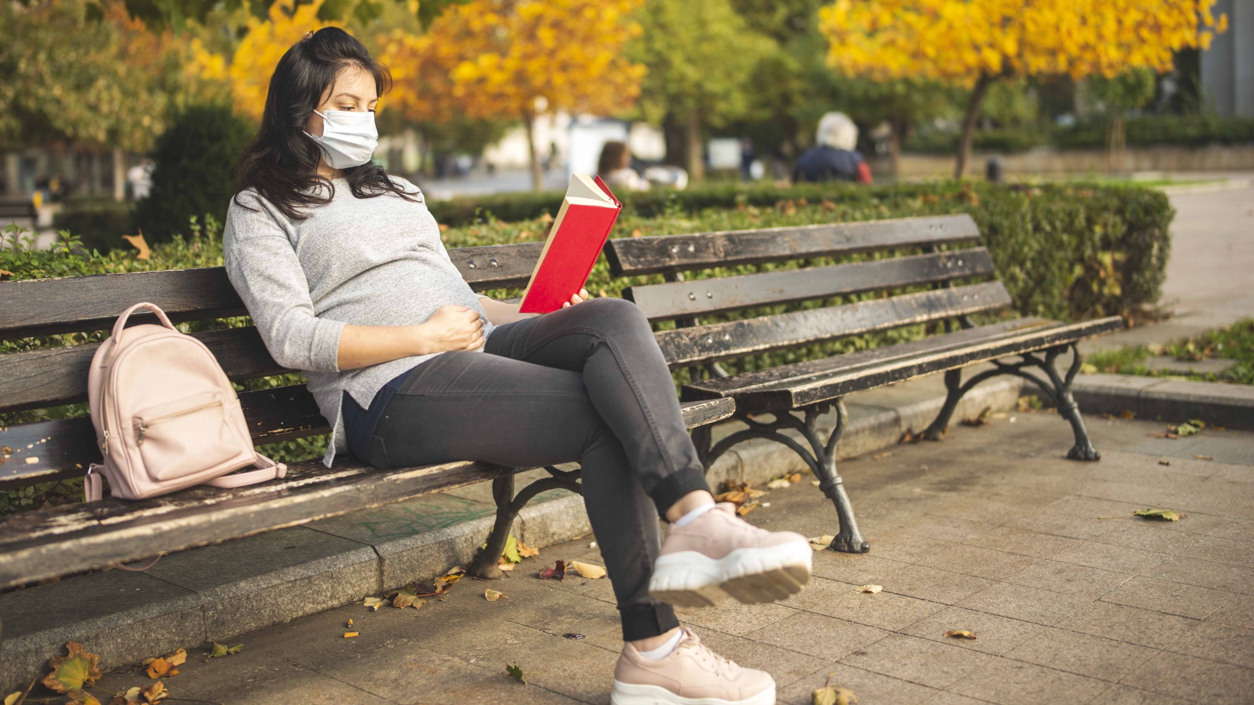 Jovem mulher grávida com máscara protetora lendo livro, sentado em um banco no parque da cidade no outono