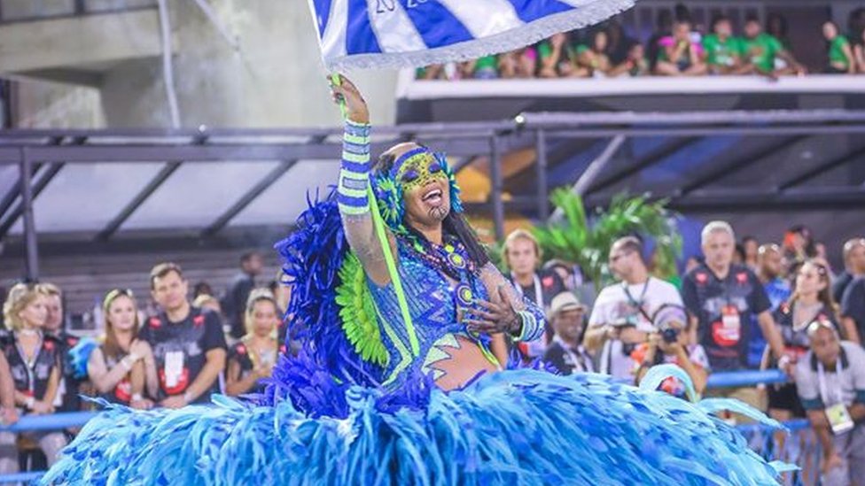 Lucinha Nobre segura bandeira enquanto desfila na Sapucaí em 2020, com fantasia azul e verde cheia de plumas