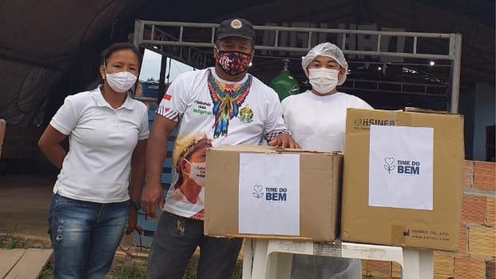 Vanda e dois homens de máscara olham para foto, em área externa, ao lado de mesca com duas caixas de papelão