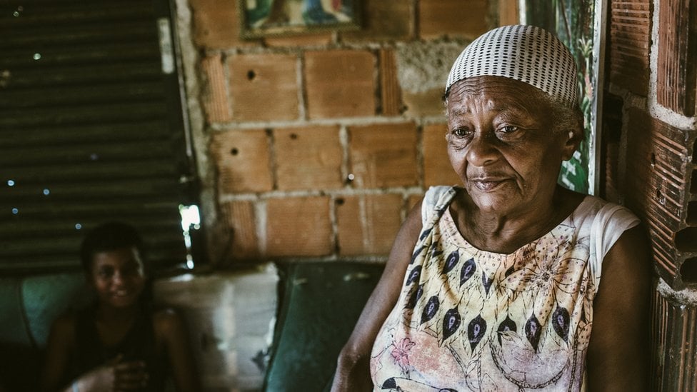 Mulher negra de lenço na cabeça com olhar triste, com menino sorridente ao fundo