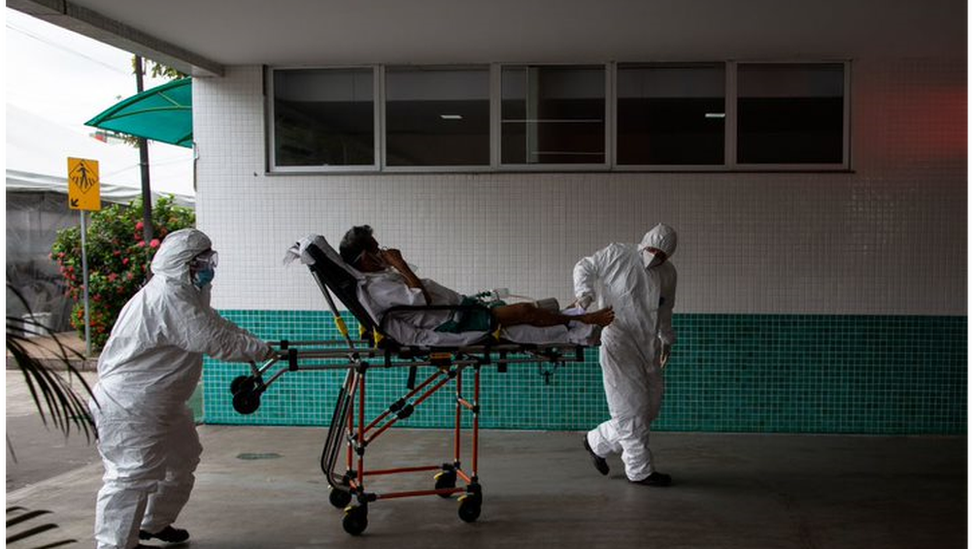Duas pessoas paramentadas carregam paciente em maca em parte externa de hospital