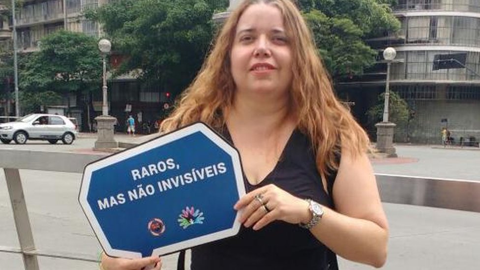 Margareth na rua, sorrindo e com placa na mão que diz: 'Raros, mas não invisíveis'