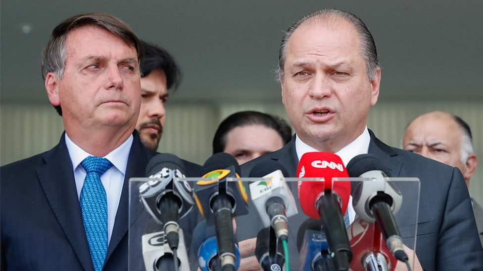 Bolsonaro olha seriamente para Barros, que fala diante de microfones de jornalistas