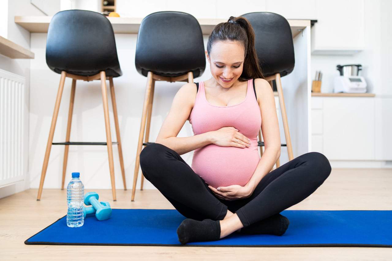 Imagem mostra uma mulher grávida, acariciando a barriga e sentada sobre um tapete de exercícios