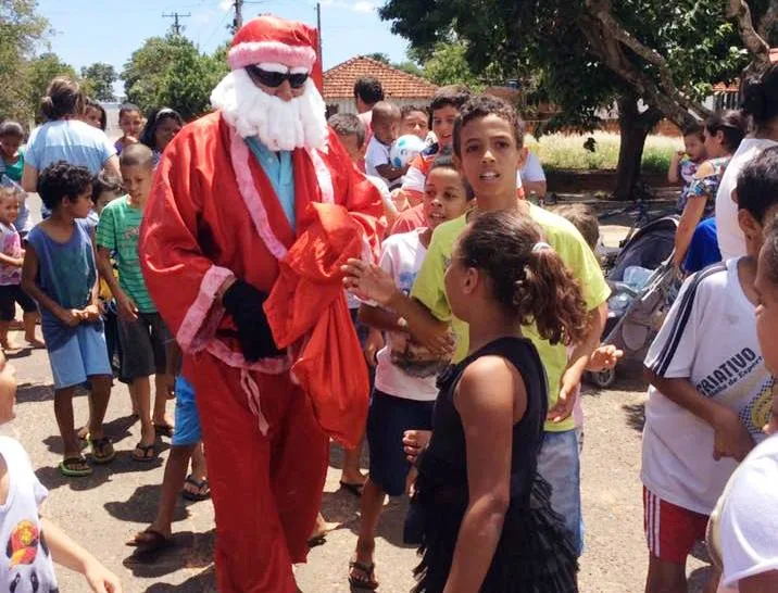 homem vestido com capuz de papai noel entrega sacolas com presentes para grupo de crianças